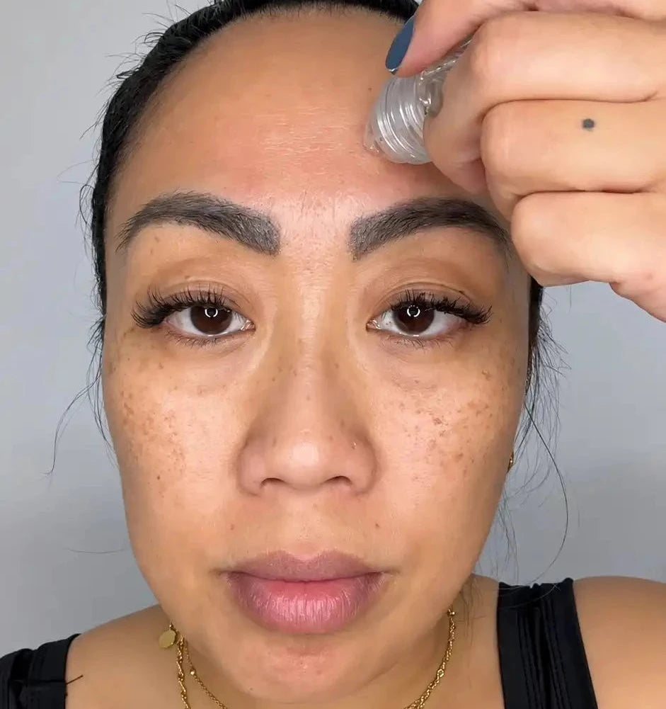 A close-up of a woman applying microinfusion to her forehead, wearing a black tank top and layered gold necklaces against a plain light gray background.