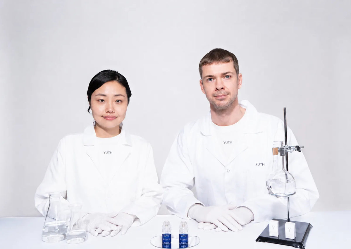 Two scientists in white lab coats and gloves sitting at a table with glassware and small blue bottles, photographed against a clean light gray studio background.
