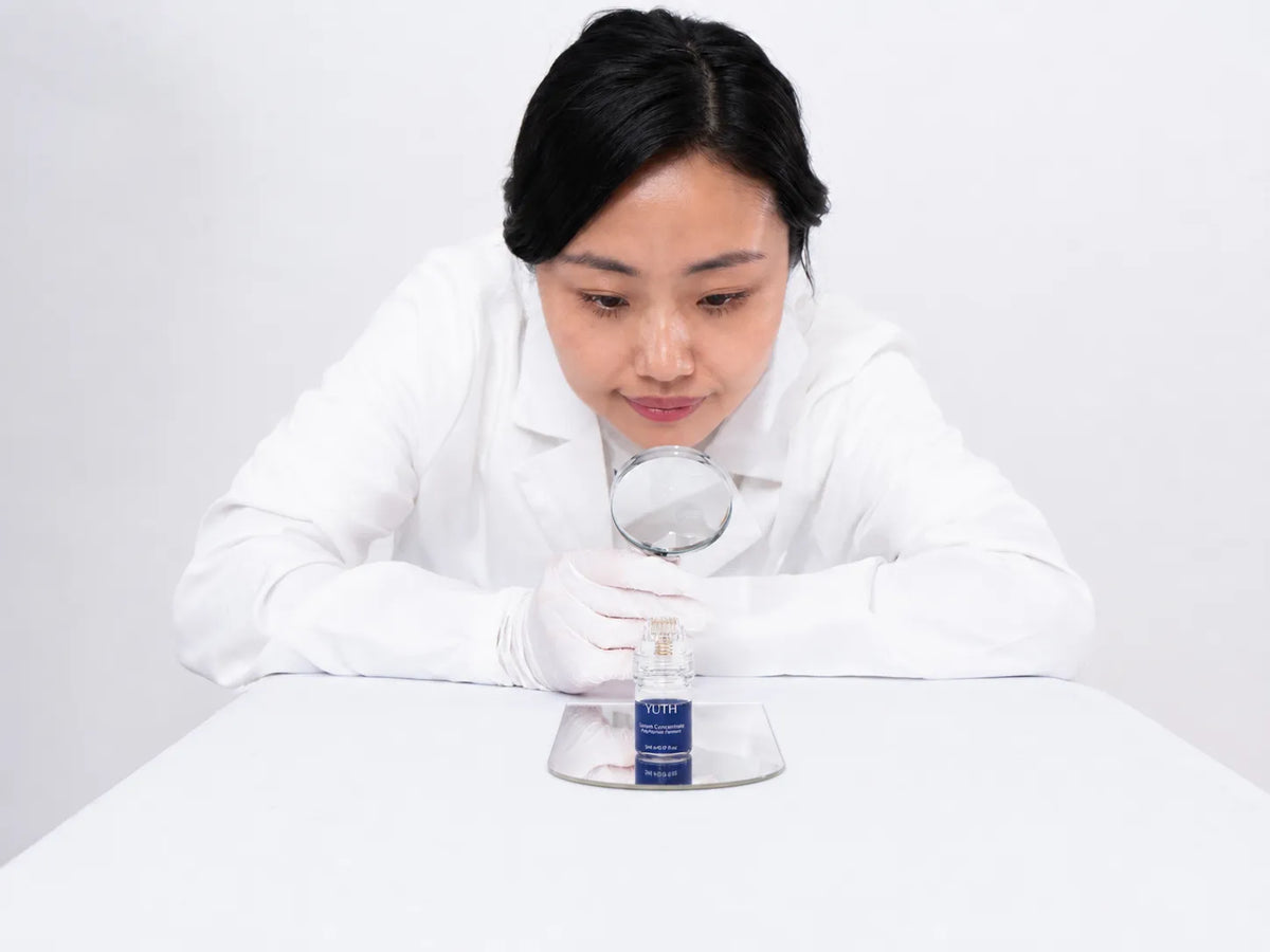 A woman in a white lab coat and gloves uses a magnifying glass to closely examine a small cosmetic bottle on a white table against a light gray background.