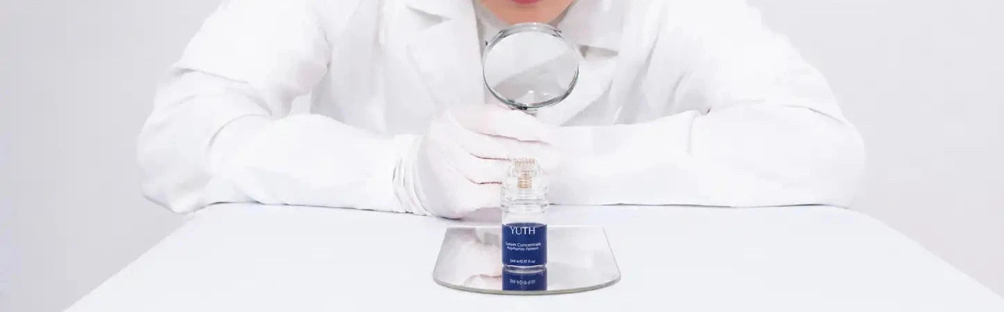 A person in a white lab coat and gloves uses a magnifying glass to examine a small bottle on a metal tray. The product appears to be a skincare or cosmetic bottle with a blue label, photographed against a clean white background.