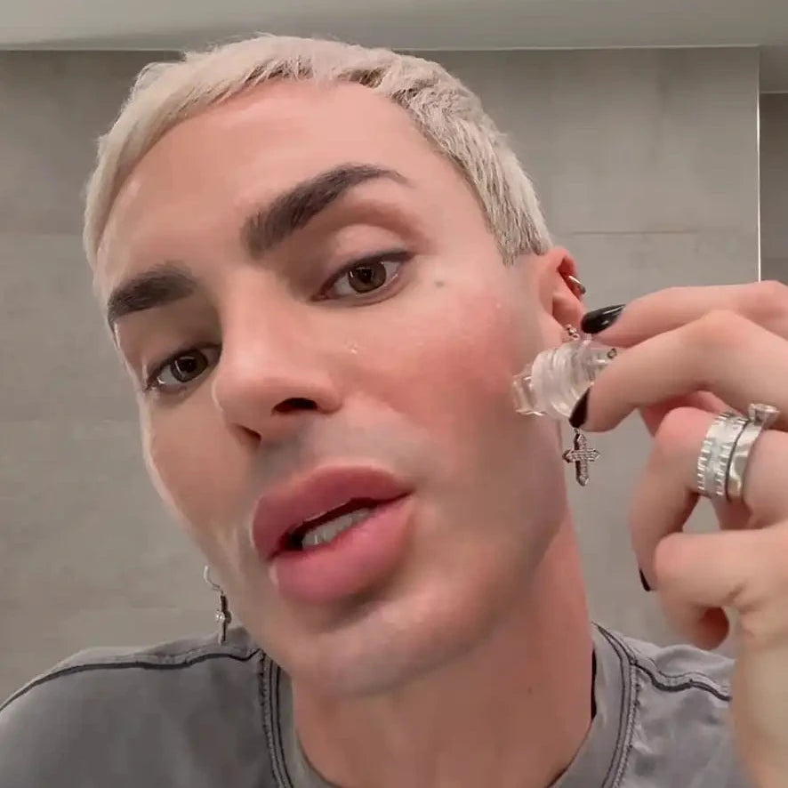 Eine Nahaufnahme eines Selfies einer Person mit platinblondem Haar und grauem T-Shirt, die ein kleines durchsichtiges Glasfläschchen neben ihr Ohr hält, mit einem Badezimmerspiegel im Hintergrund und einer Kommentarblase am unteren Rand.