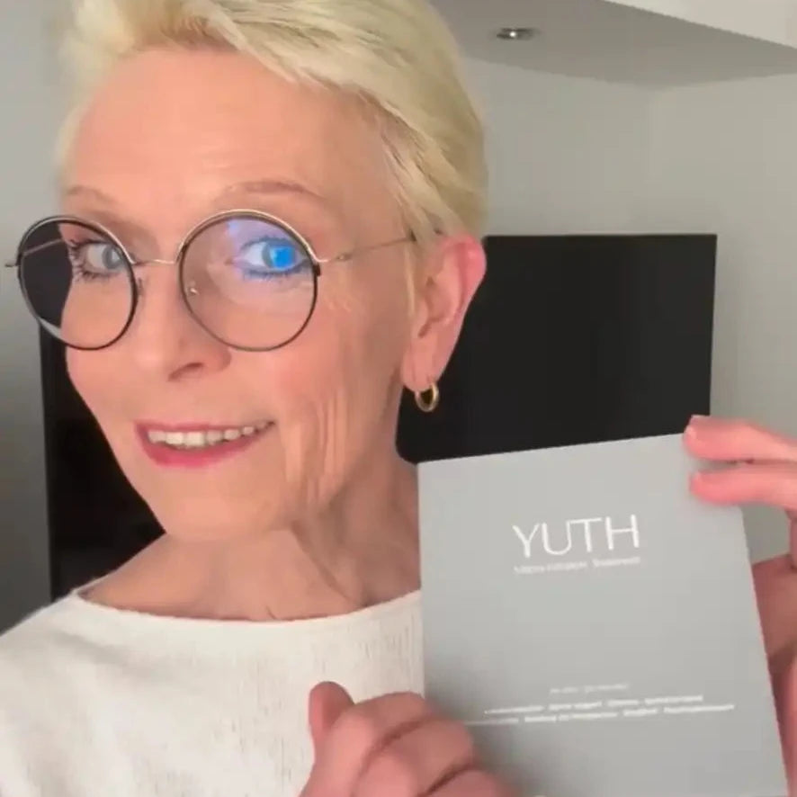 Eine Frau mit kurzem blondem Haar und einer runden Brille hält eine graue YUTH-Mikroinfusions-Hautpflegebox hoch und lächelt in einer Innenaufnahme in die Kamera.